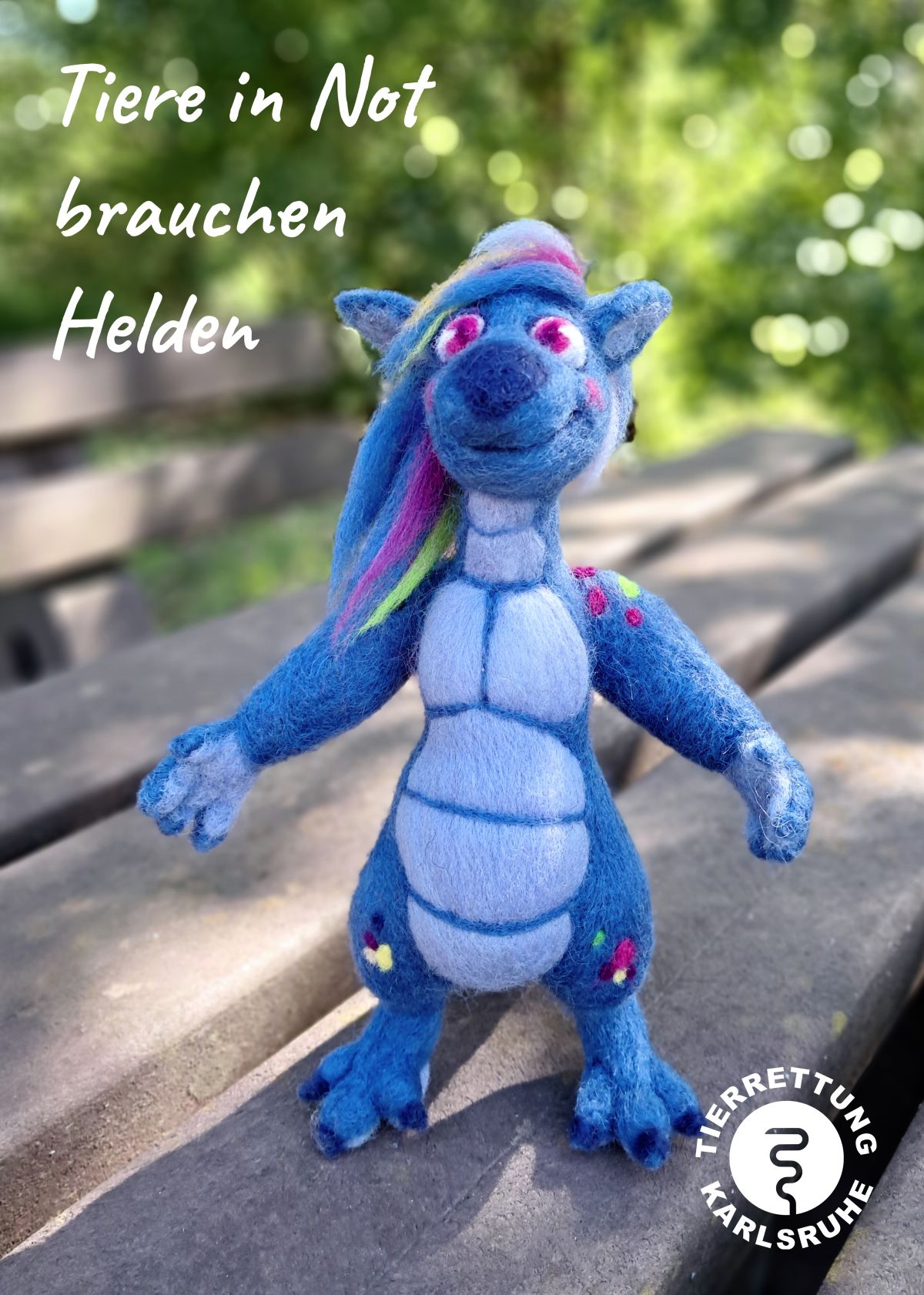 Eine kleine blaue Drachenfigur aus Filz sitzt auf einem Tisch und im Hintergrund erkennt man verschwommen grüne Pflanzen. Oben links ist ein weißer Schriftzug Tiere in Not brauchen Helden und unten rechts ein weißes rundes Logo mit Kreis und stilisierter weißer Schlange und Schriftzug Tierrettung Karlsruhe